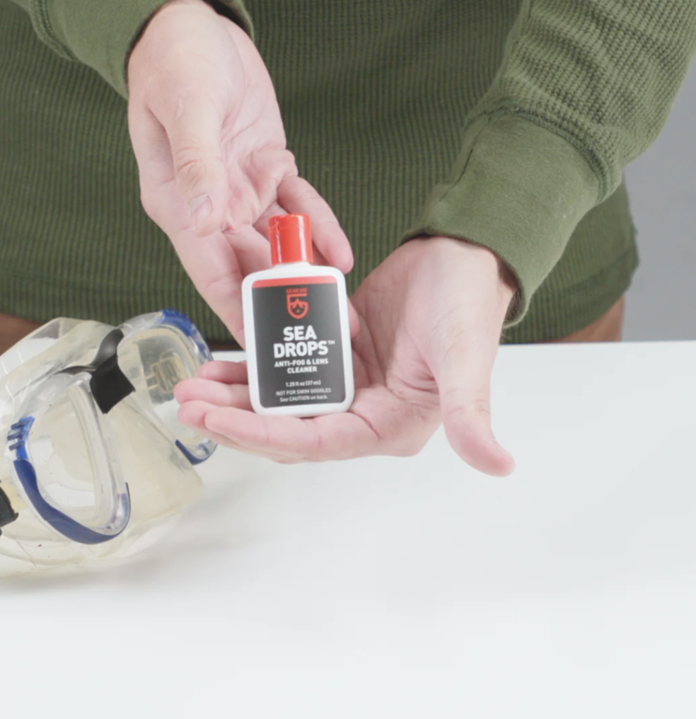 A person holding a bottle of Gear Aid Sea Drops in front of a dive mask, demonstrating its use as an anti-fog solution for glass scuba and prescription dive masks.